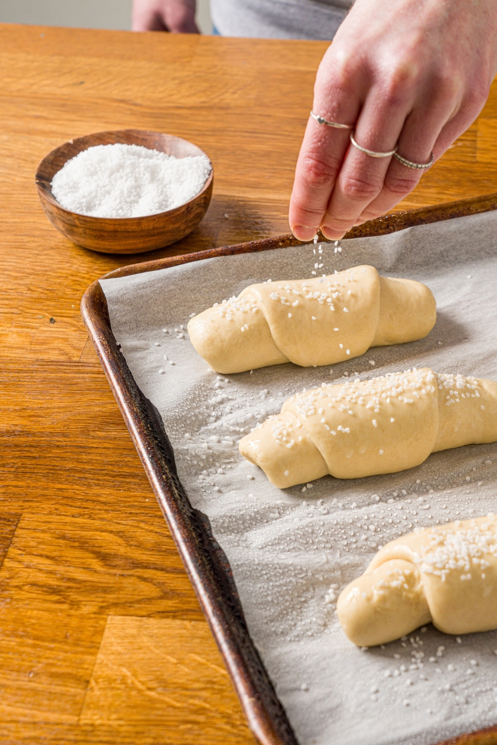 A baking sheet lined with parchment paper with rolled salt bread dough pieces. The dough is being sprinkled with salt. The sheet is on a wooden counter with a small bowl of salt.