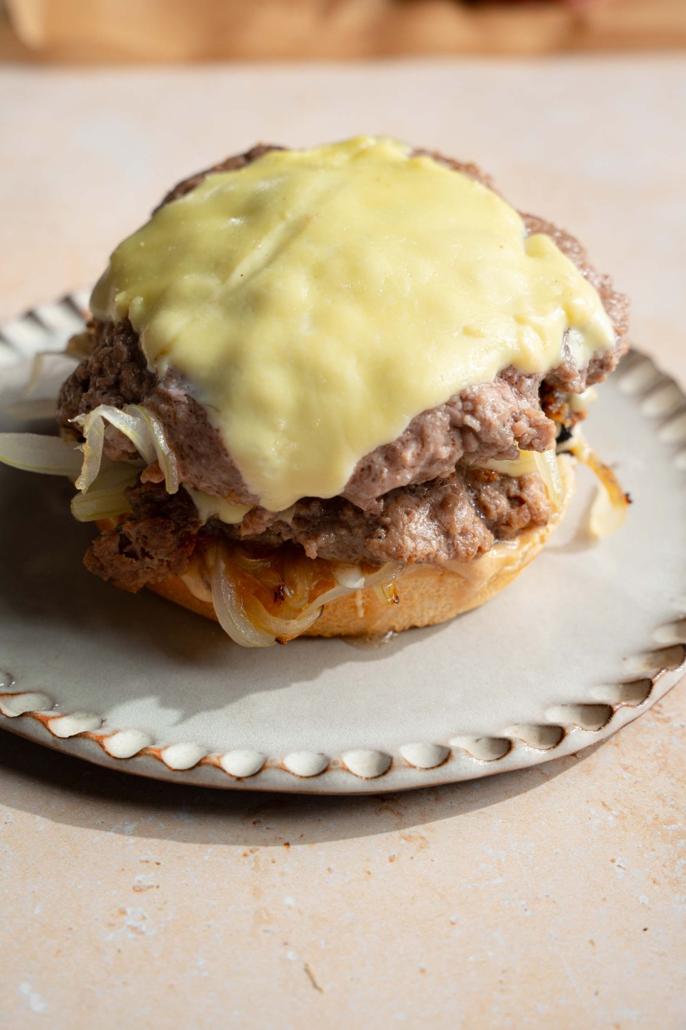 A white plate with an open smash burger with onions and cheese. The plate is on a tan counter.