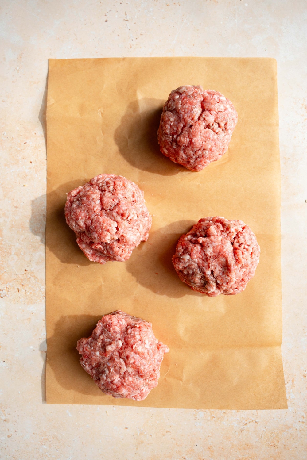 Four beef patties shaped in a ball. The balls are on a piece of parchment paper on a tan counter.