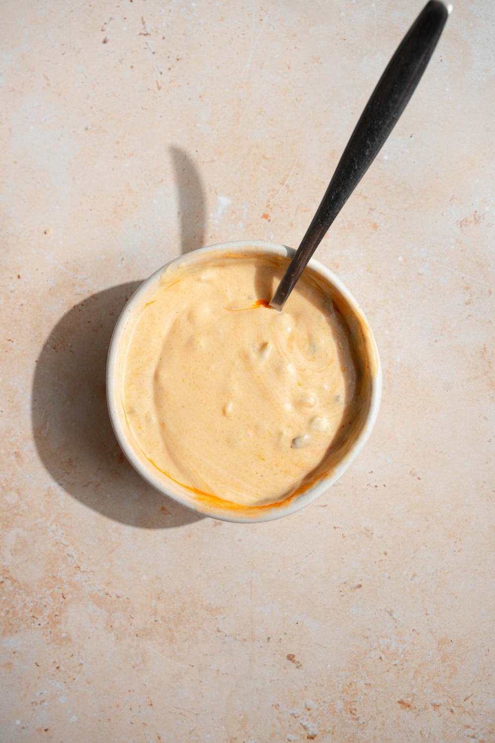 A small bowl with a spoon mixing ingredients to make smash burger sauce. The bowl is on a tan counter.