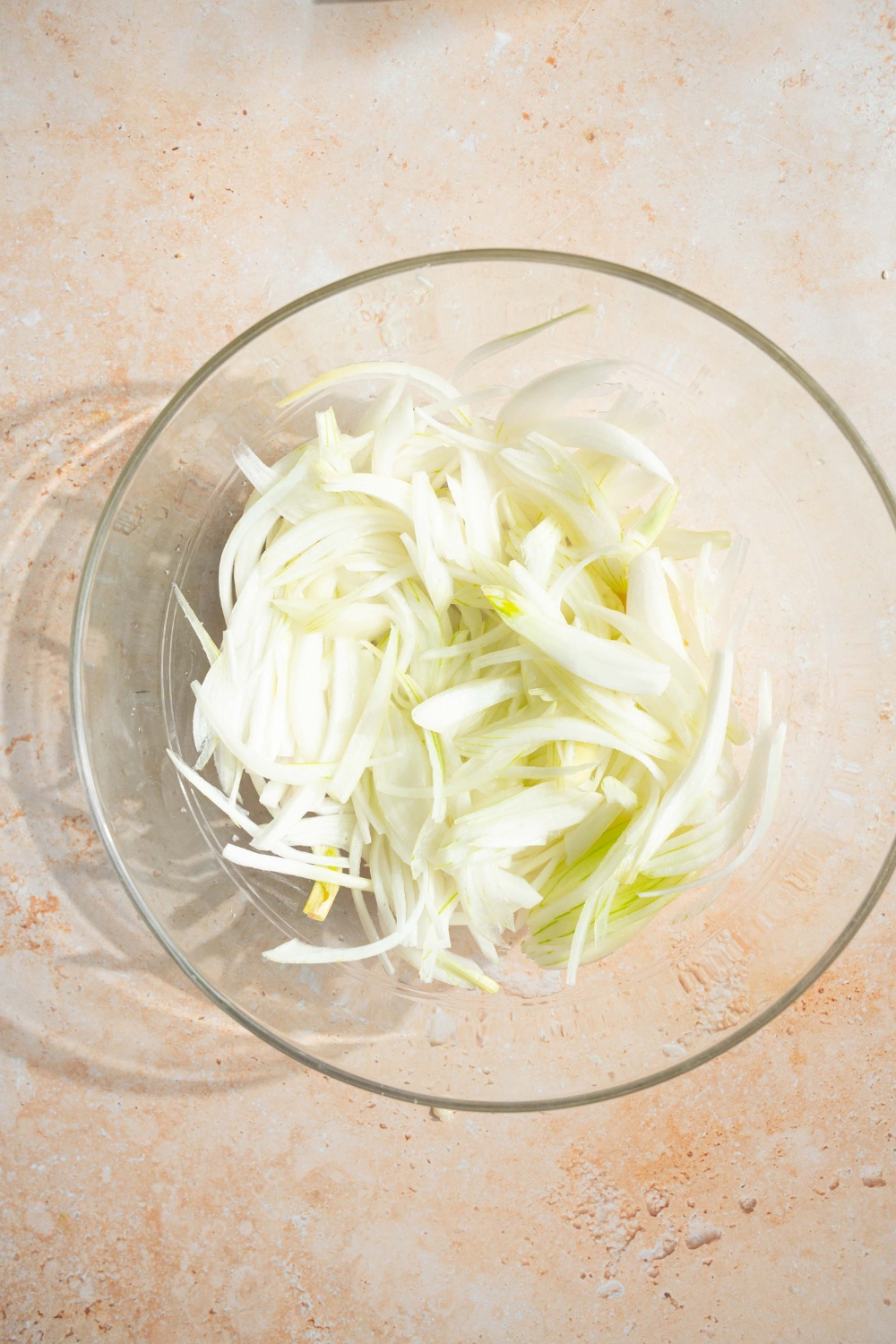 A glass bowl with sliced onions. The bowl is on a tan counter.