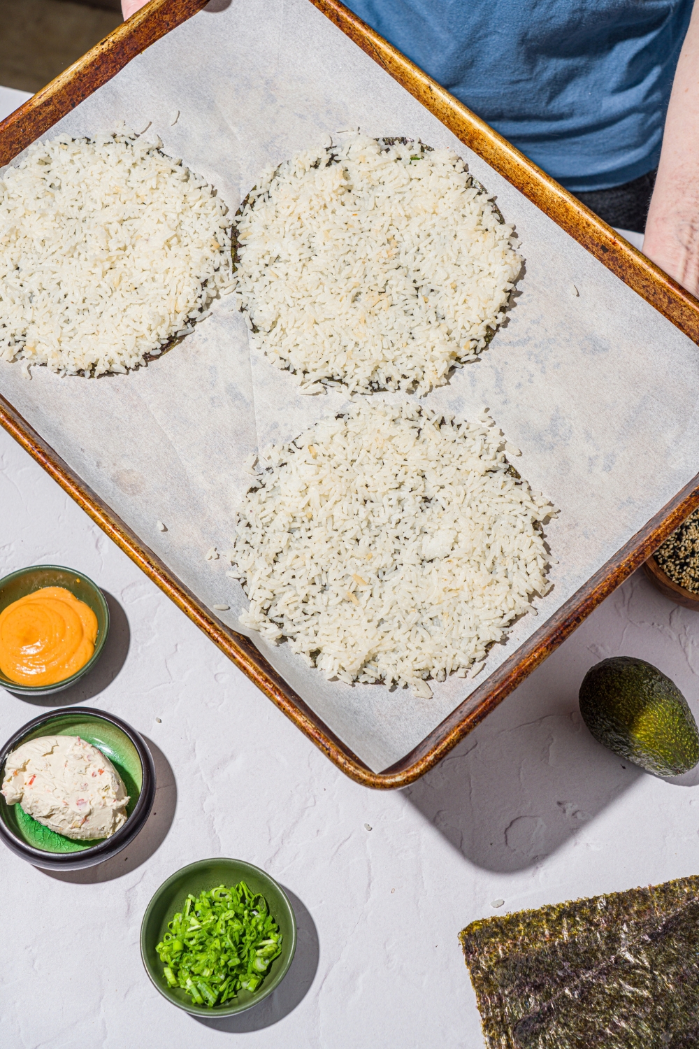A baking sheet lined with parchment paper with three nori sheets cut into circles with sushi rice on top. There is a white counter with small bowls of ingredients.