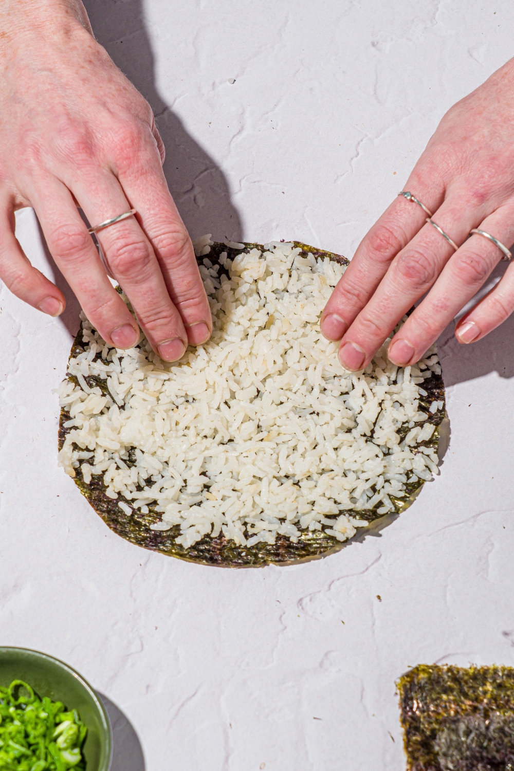 A nori sheet cut into a circle with a pair of hands spreading sushi rice over the sheet. The sheet is on a white counter.