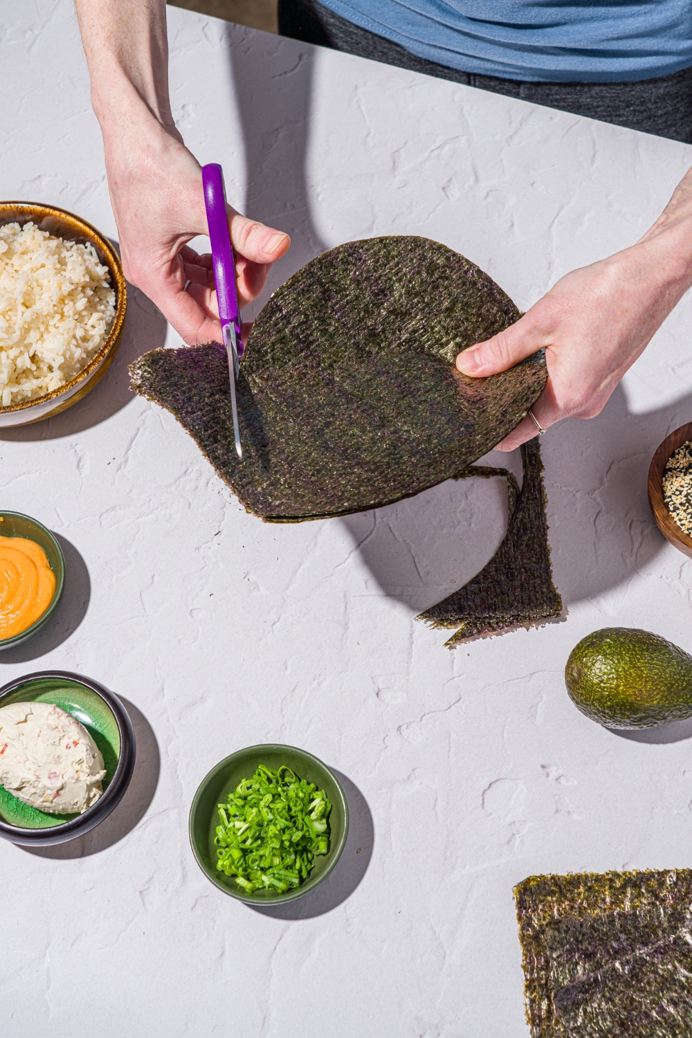 A pair of hands cutting nori sheets into a circular shape with scissors. There is a white counter with several bowls of ingredients.