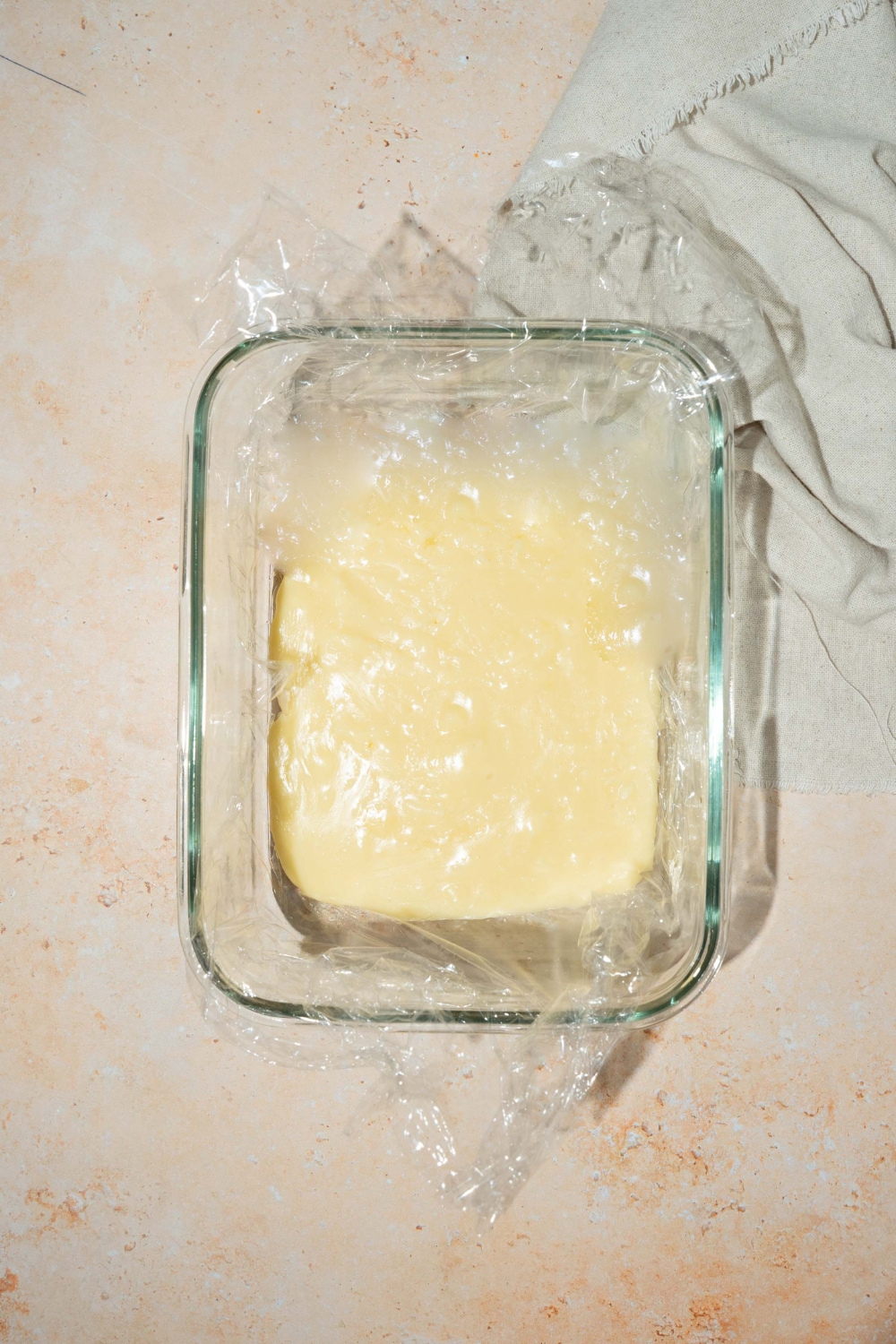 A glass baking dish with vanilla fudge mixture covered with plastic wrap. The dish is on a tan counter with a white cloth napkin.