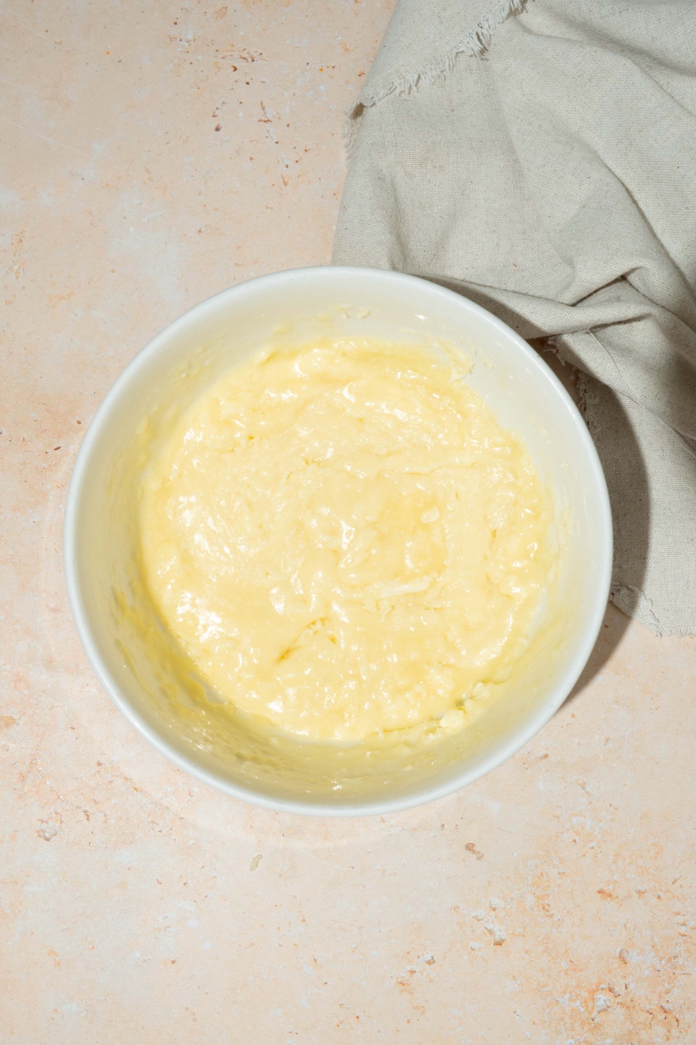 A white bowl with melted white chocolate and condensed milk mixture. The bowl is on a tan counter with a white cloth napkin.