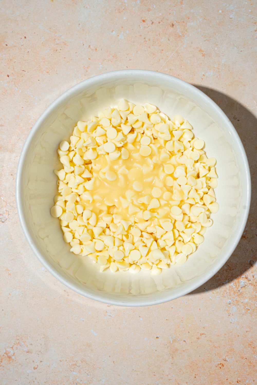 A white bowl with white chocolate pieces and condensed milk. The bowl is on a tan counter.