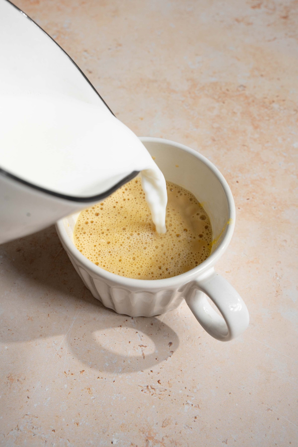 A white mug with milk being poured into the mixture to make Vietnamese egg coffee. The mug is on a tan counter.