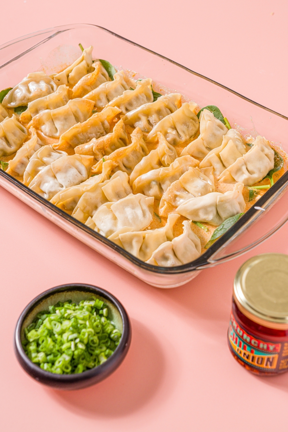 A glass baking dish with uncooked Trader Joe's chili crisp dumpling bake. The dish is on a pink counter with a small bowl of sliced green onions and jar of Trader Joe's chili onion crunch.