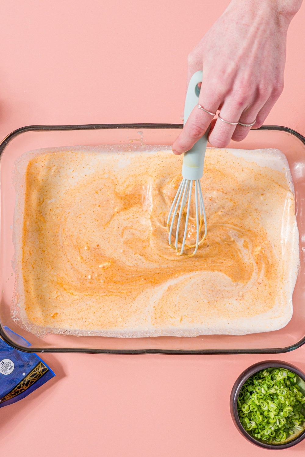 A glass baking dish with a whisk mixing coconut milk, soyaki, thai red curry sauce, and garlic. The dish is on a pink counter with a small bowl of sliced green onions.