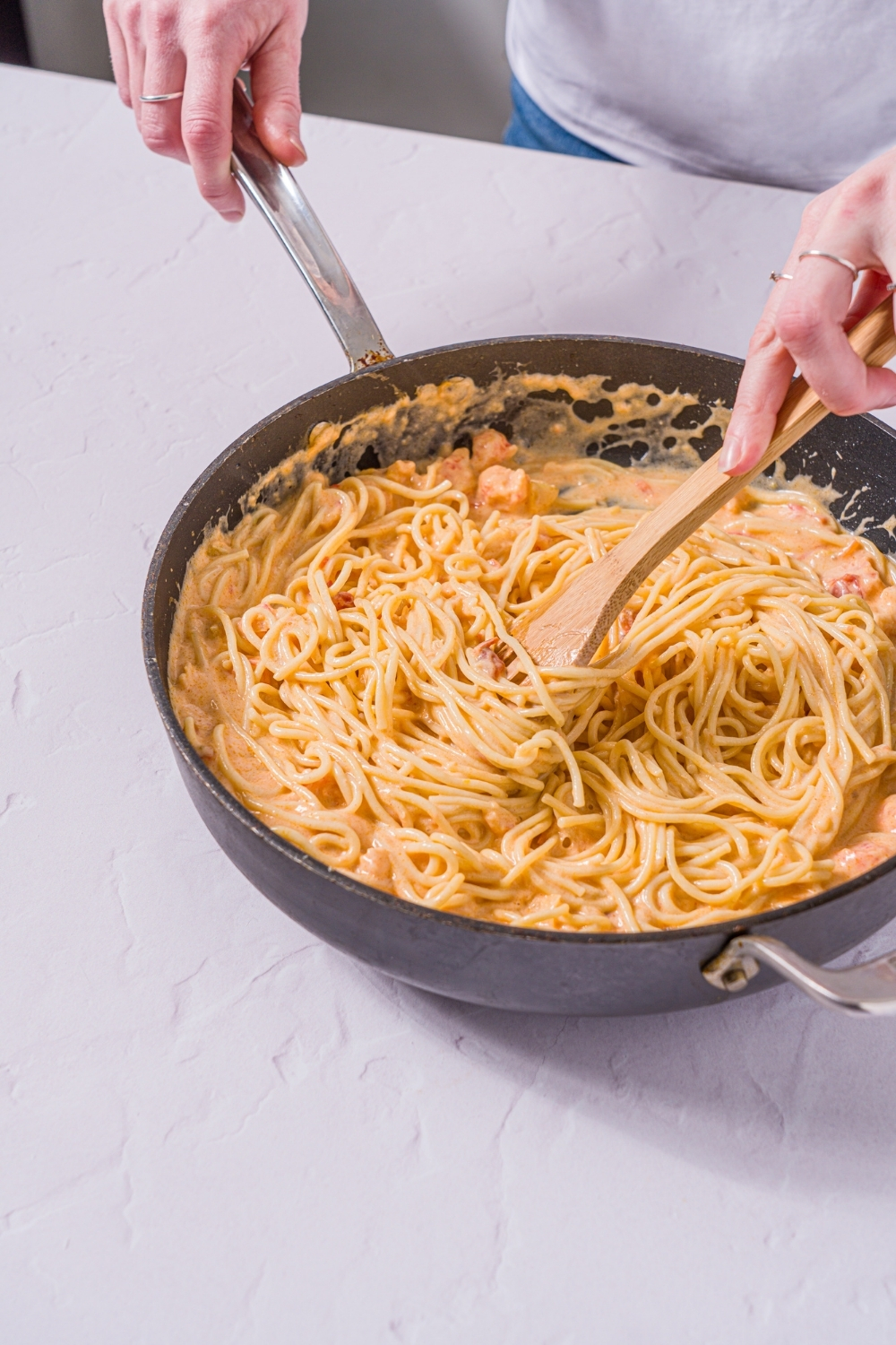 A skillet with spaghetti tossed in a Trader Joe's lobster bisque sauce. A wooden spoon is stirring the pasta. The skillet is on a marble counter.