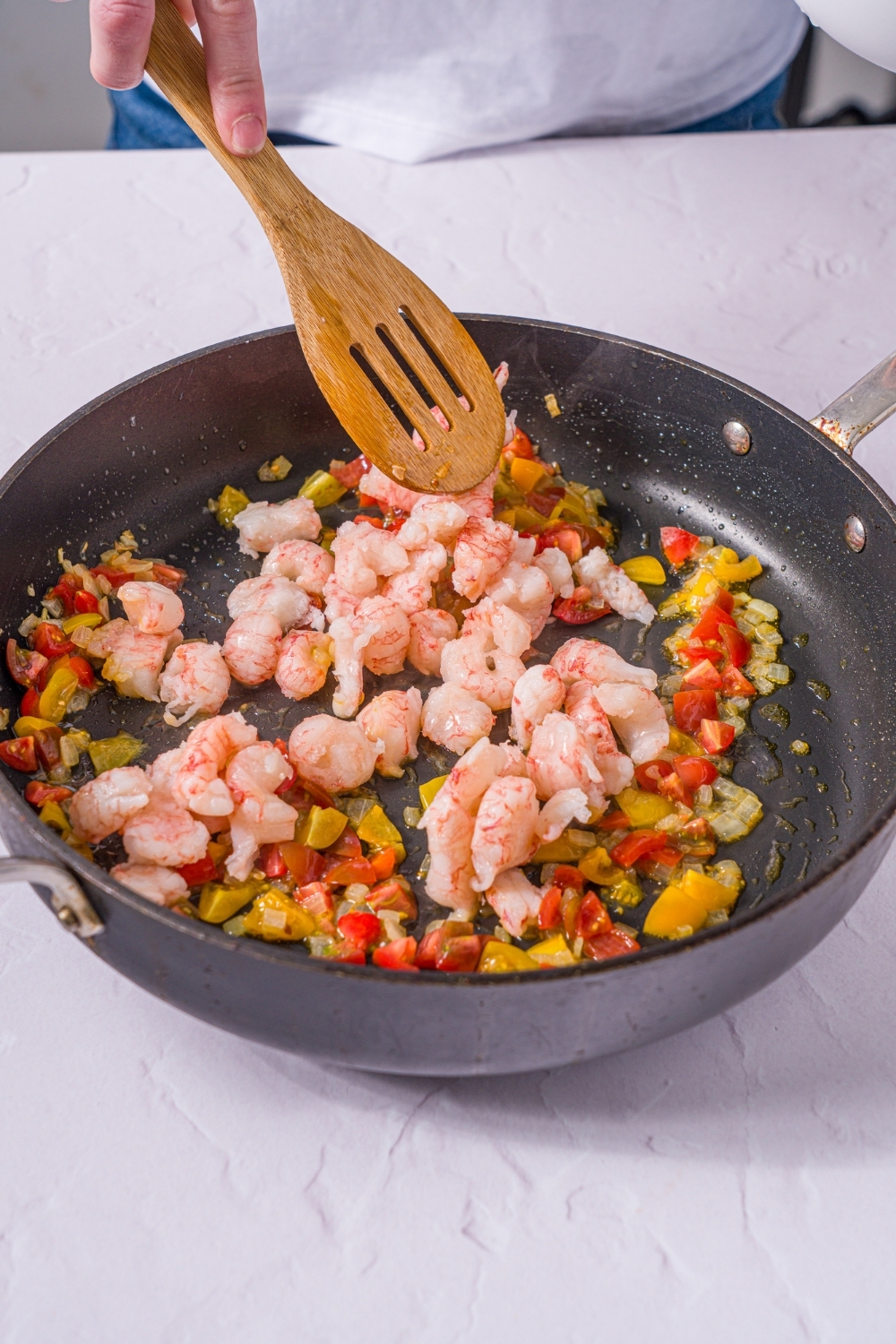 A skillet with onions, tomatoes, and shrimp sautéing in oil. A wooden spoon is stirring the mixture. The skillet is on a marble counter.