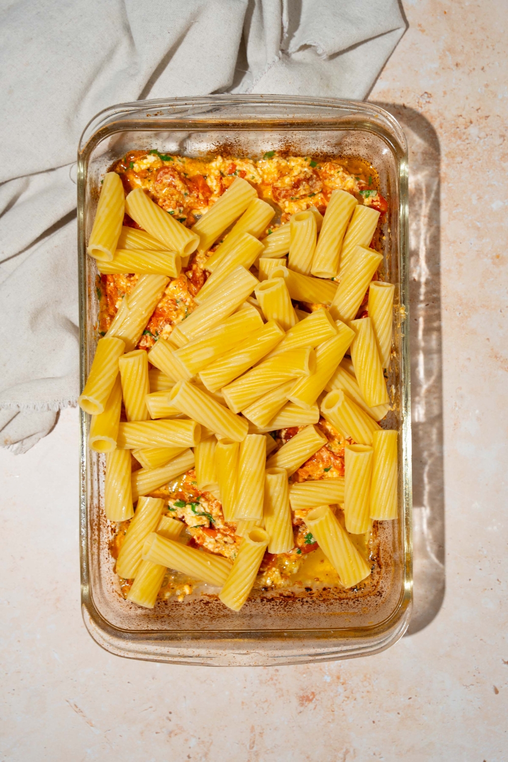 A glass baking dish with rigatoni pasta over a feta cheese and cherry tomatoes mixture. The dish is on a tan counter with a white cloth napkin.