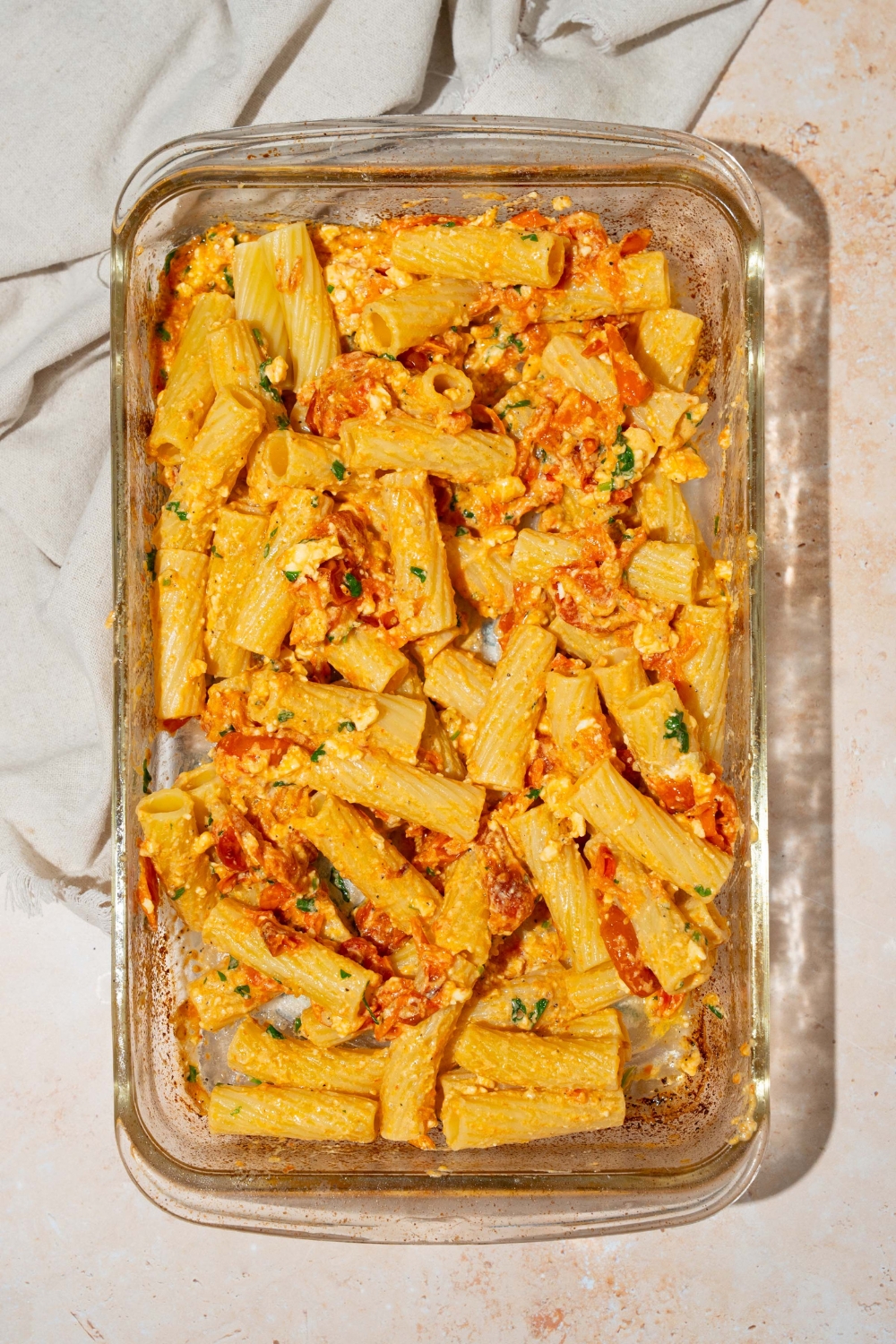 A glass baking dish with rigatoni pasta tossed in a feta cheese and cherry tomatoes mixture. The dish is on a tan counter with a white cloth napkin.