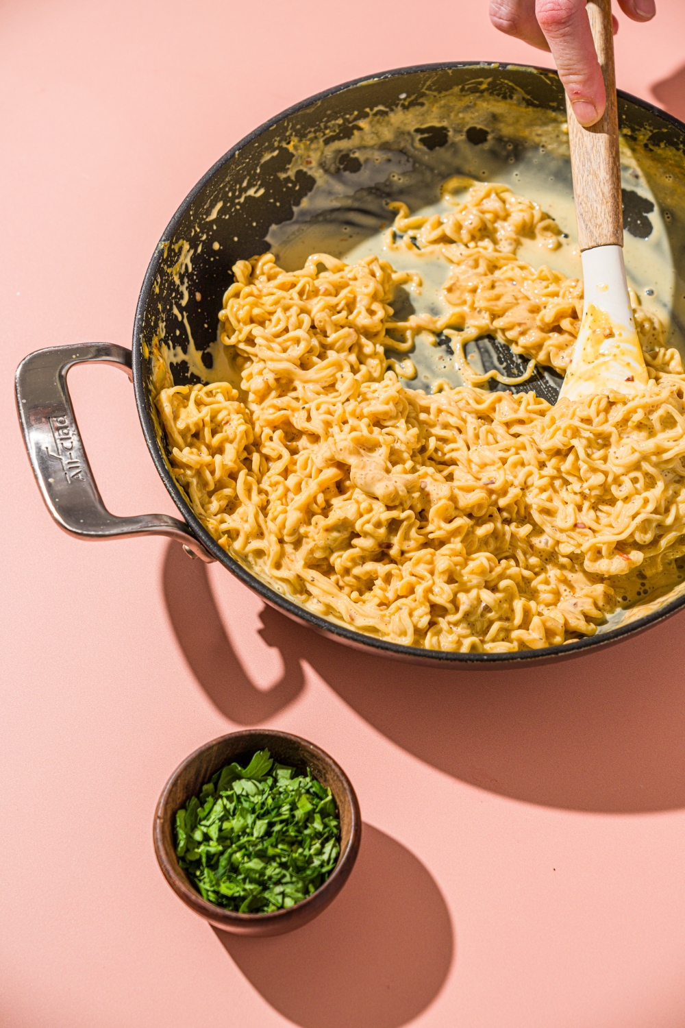 A skillet with a spatula stirring cheesy maggi noodles. The skillet is on a pink counter with a small bowl of cilantro.