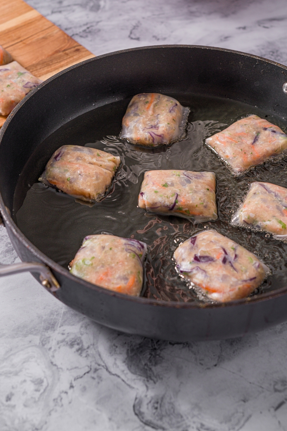 A skillet with several chicken and shrimp rice paper dumplings frying in oil. The skillet is on a marble counter.