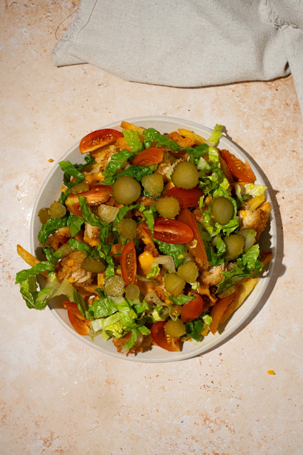 A white plate with chicken burger bowl loaded fries with fries topped with crispy chicken, lettuce, shredded cheese, tomatoes, and pickles. The plate is on a tan counter with a white cloth napkin.