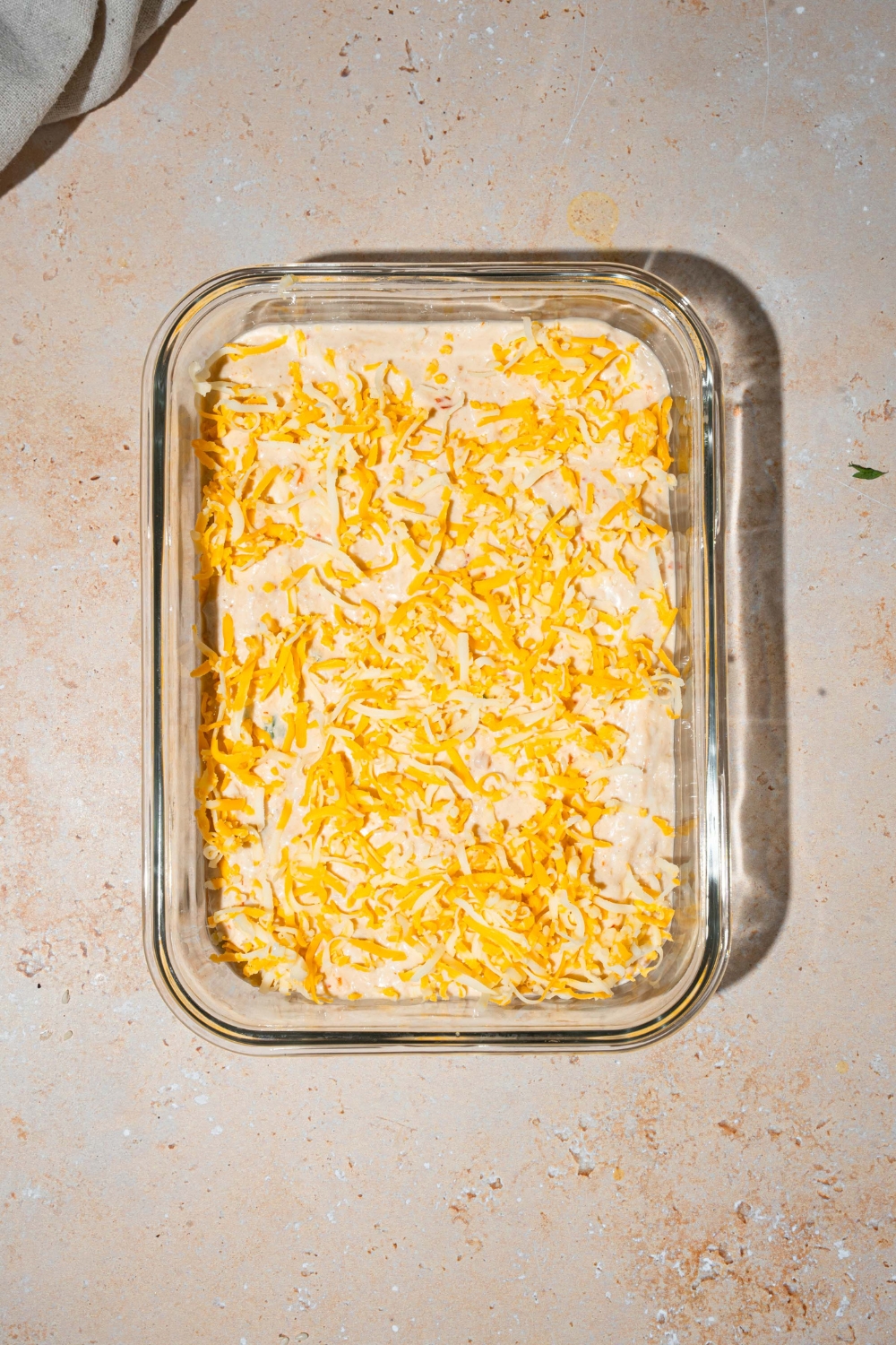 A glass baking dish with unbaked crab rangoon mix topped with shredded cheese. The dish is on a tan counter.