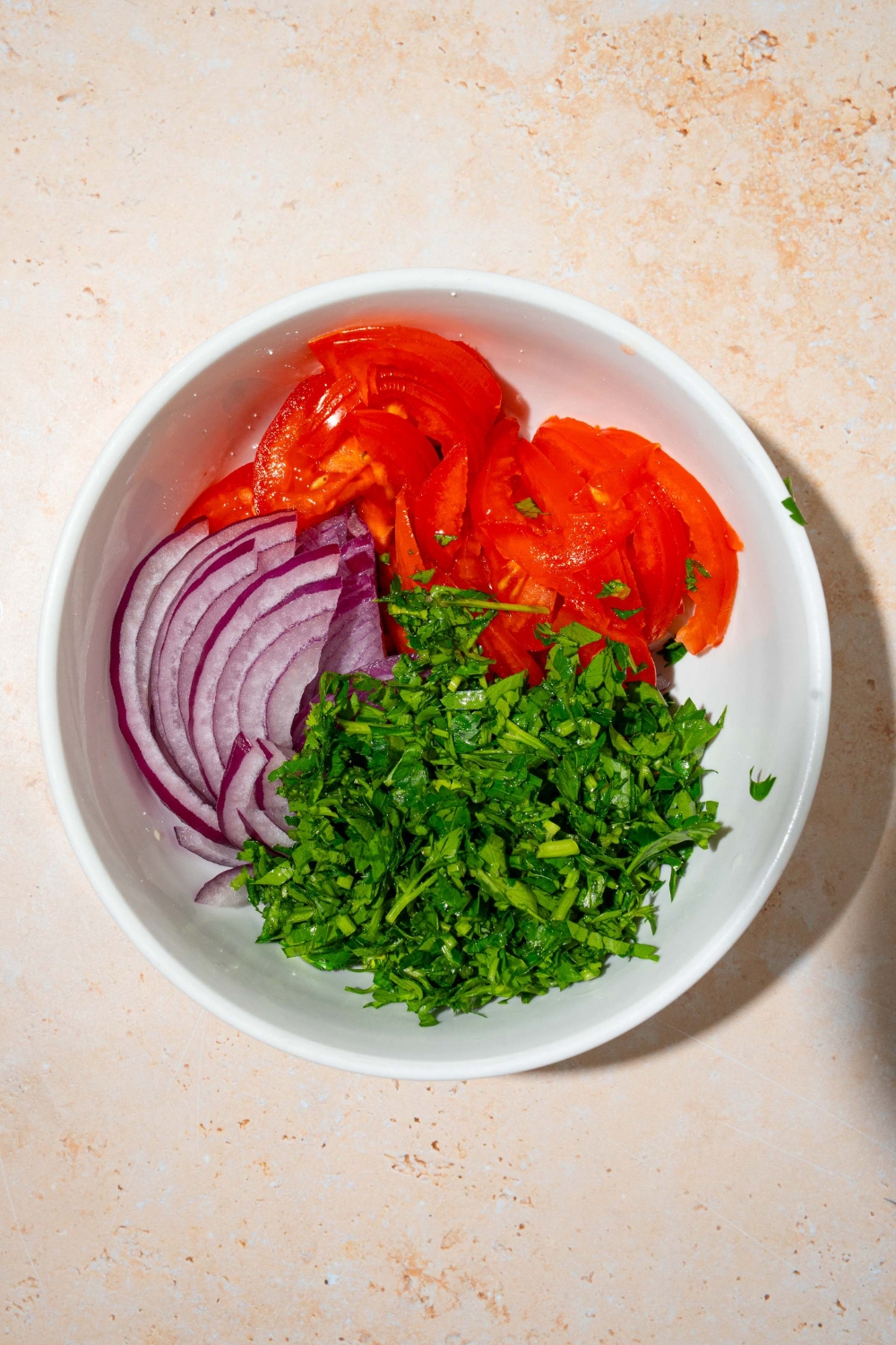 A white bowl with sliced red onion, sliced tomatoes, and fresh parsley. The bowl is on a tan counter.