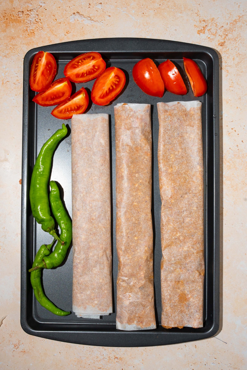 A baking sheet with three doner kebab meat logs wrapped in parchment paper with green peppers and sliced tomatoes. The sheet is on a tan counter.