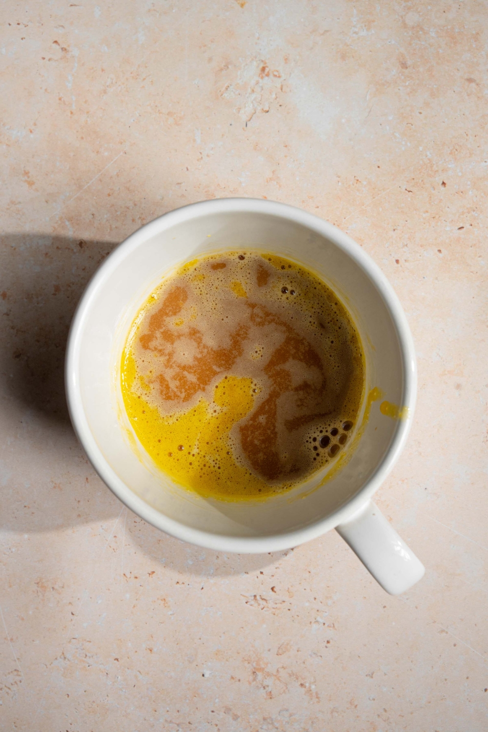 A white mug with a whisked yolk and coffee mixture. The mug is on a tan counter.