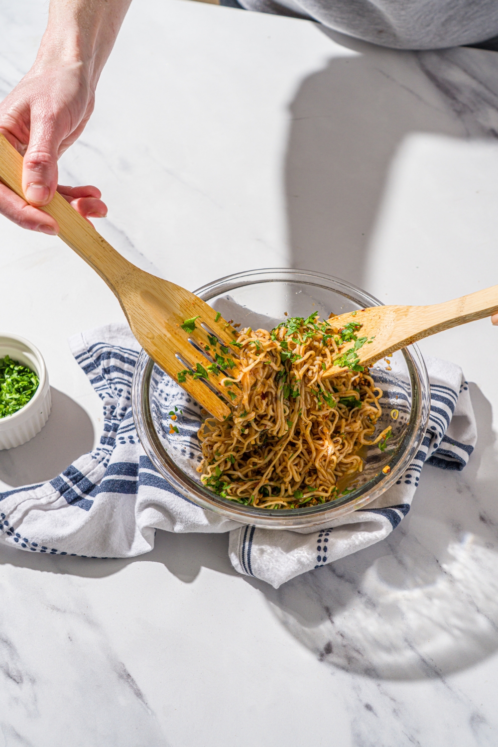A glass bowl with two wooden spoons mixing rice noodles with chili oil sauce. The bowl is on a marble counter with a white cloth napkin.