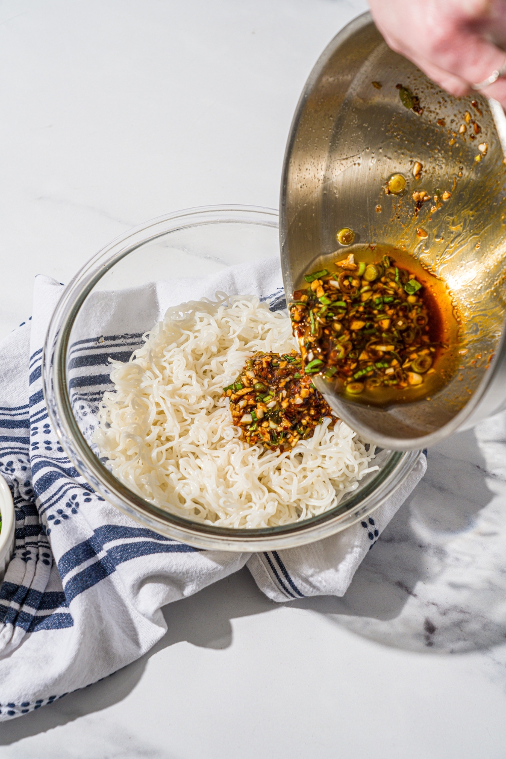 A glass bowl with cooked rice noodles. A bowl is pouring garlic chili oil into the noodles. The bowl is on a marble counter with a white cloth napkin.