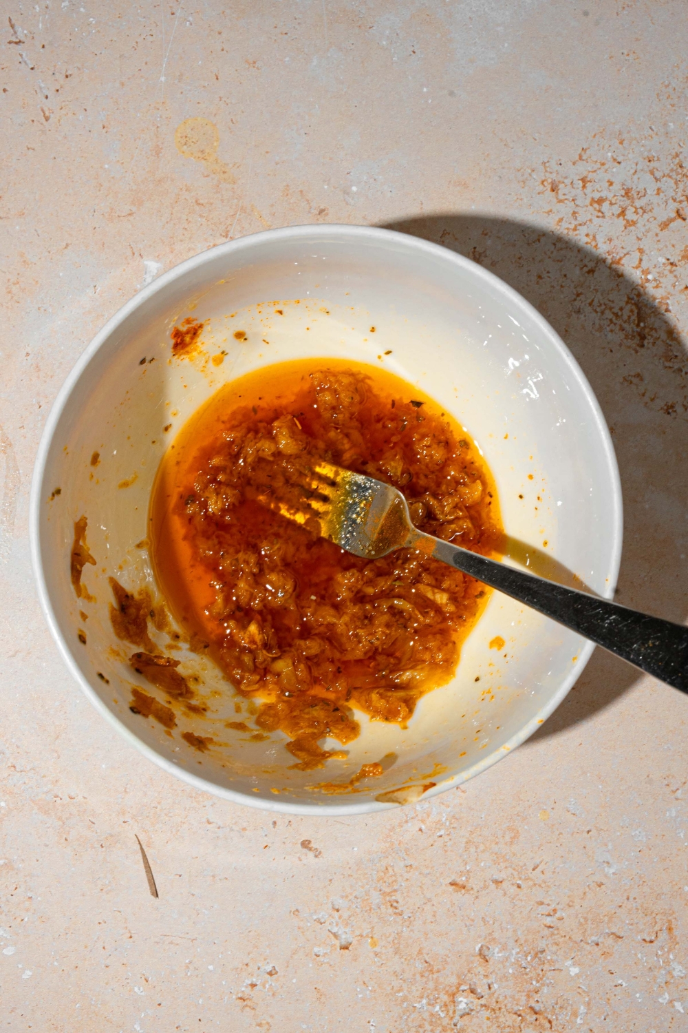 A white bowl with a fork mashing the garlic with paprika and oil. The bowl is on a tan counter.