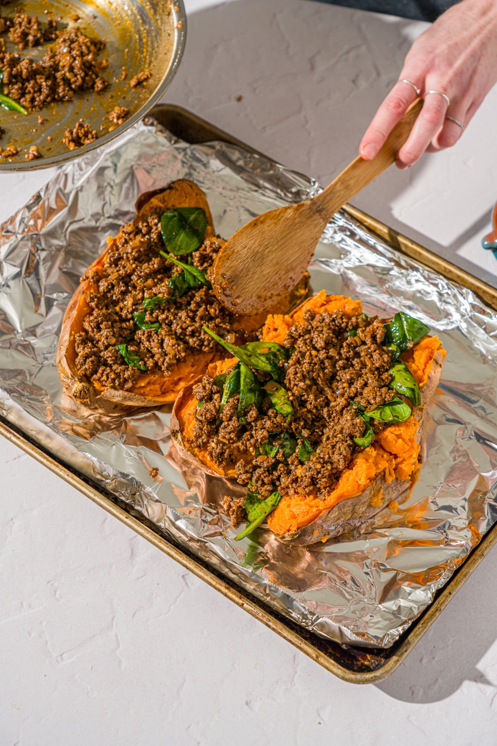 A baking sheet lined with foil with two cooked sweet potatoes sliced open. A spoon is stuffing a beef mixture into the potatoes. The sheet is on a white counter.