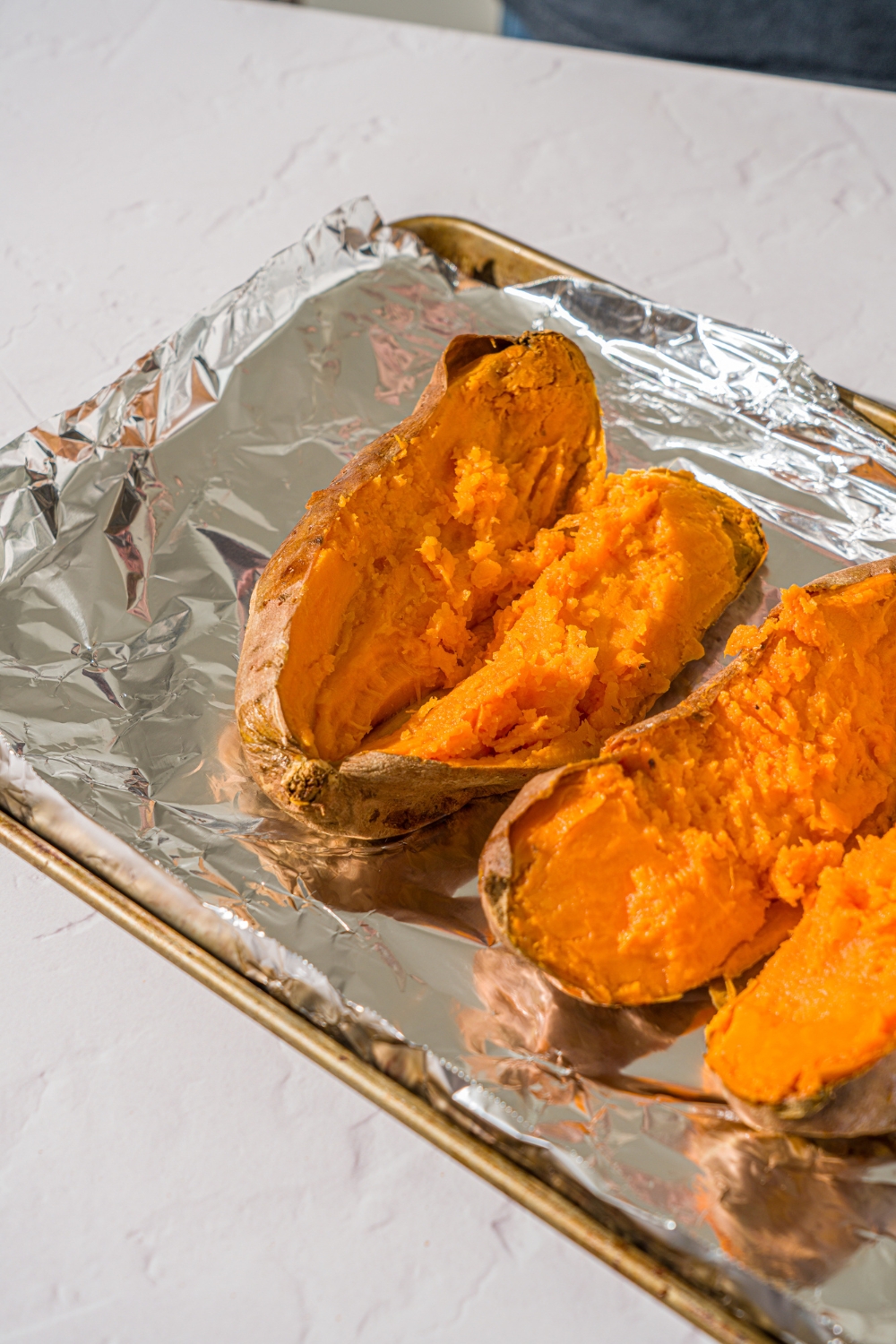 A baking sheet lined with foil with two baked sweet potatoes sliced open. The sheet is on a white counter.