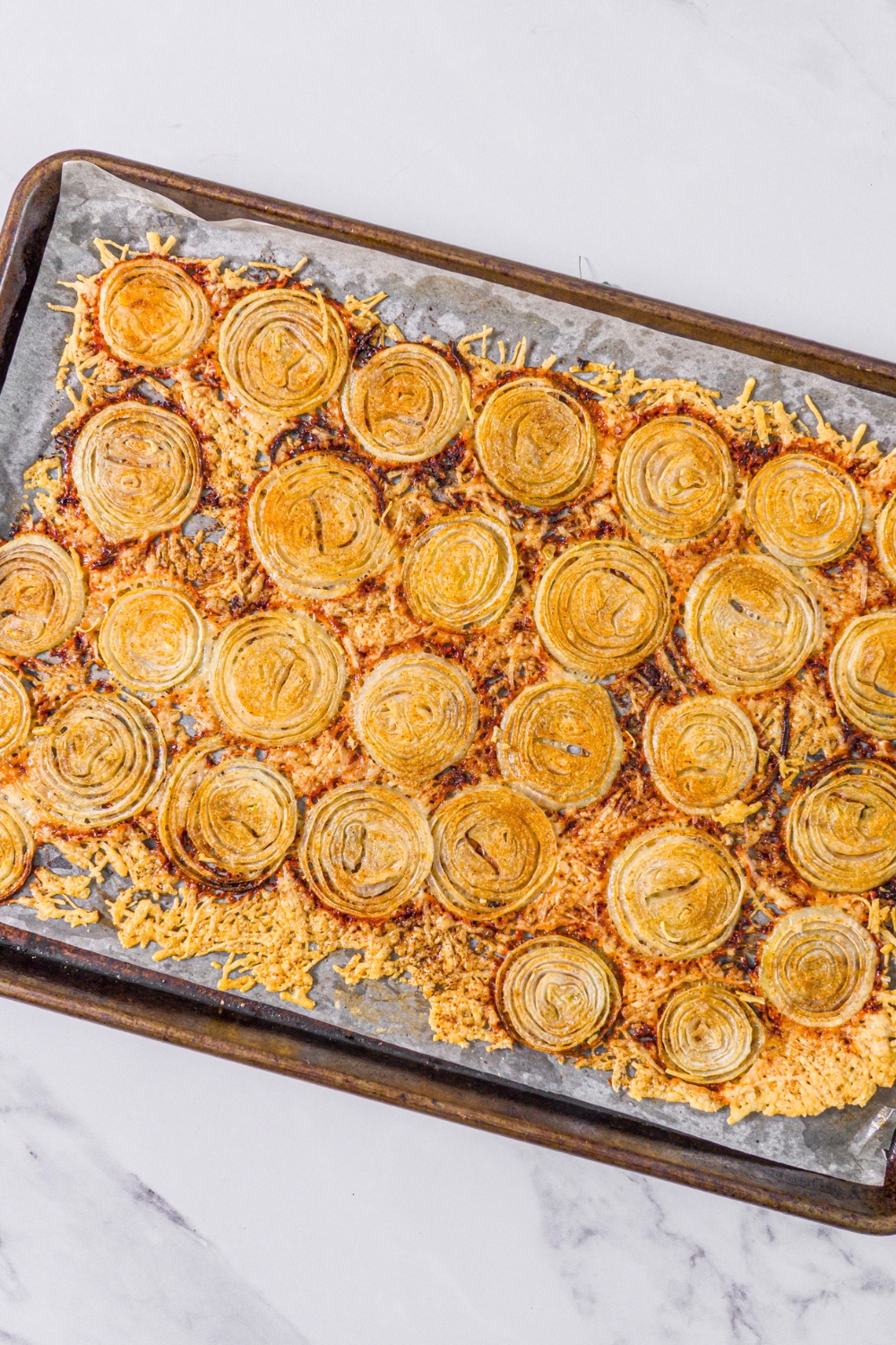 A baking sheet lined with parchment paper with baked parmesan onion chips. The sheet is on a marble counter.
