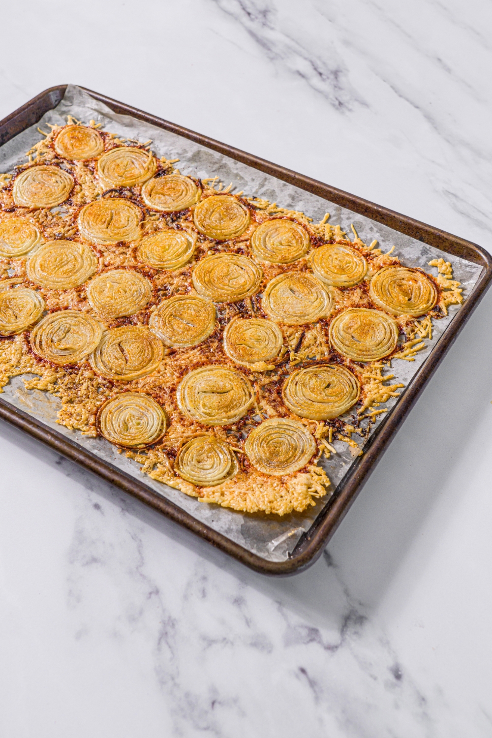 A baking sheet lined with parchment paper with baked parmesan onion chips. The sheet is on a marble counter.