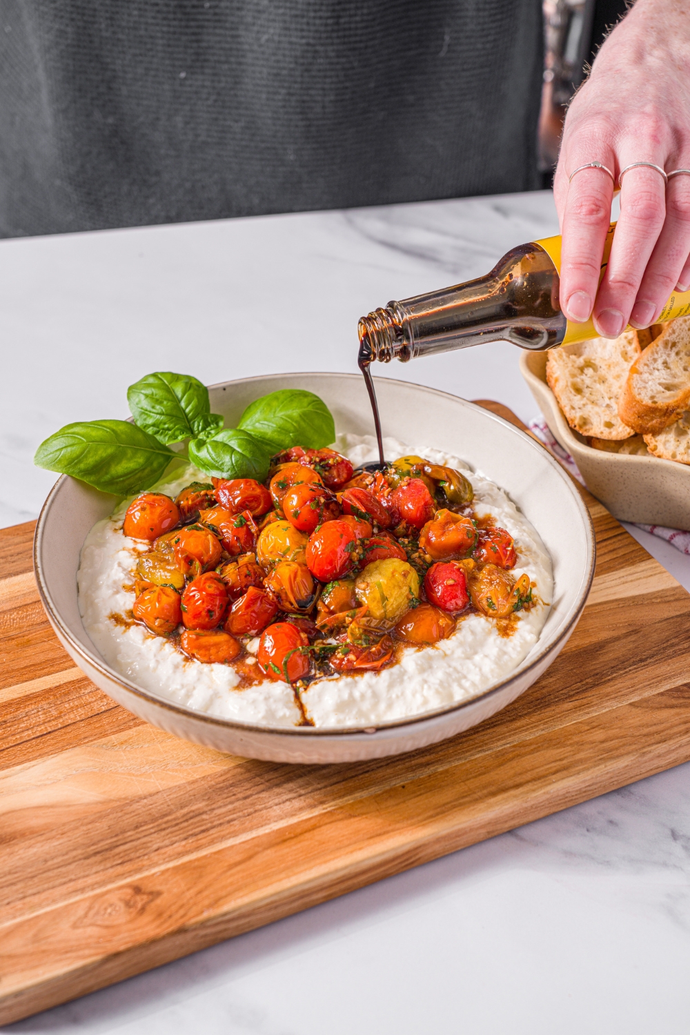 A white bowl with roasted tomato burrata dip with balsamic being drizzled over the dip. The bowl is garnished with fresh basil. The bowl is on a wooden board on a marble counter.