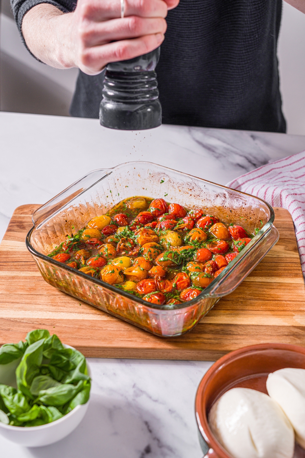 A glass baking dish with a tomato mixture with olive oil, balsamic vinegar, and seasonings. A pepper mill is grinding pepper over the dish. The dish is on a wooden board. The board is on a marble counter.