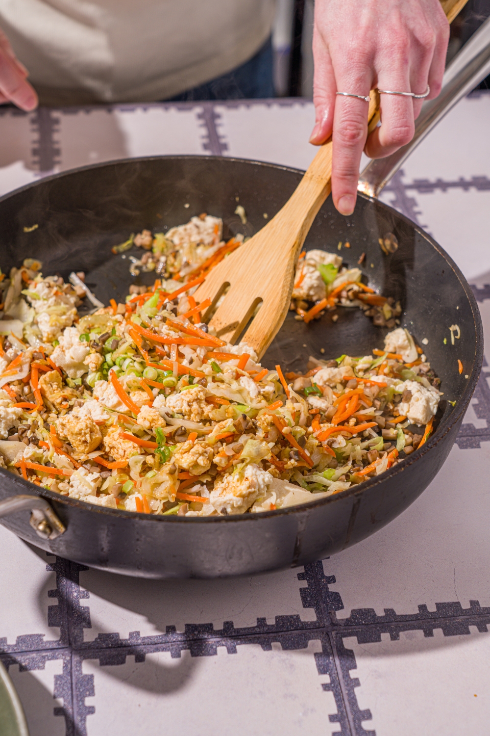 A skillet with a wooden spoon cooking a vegetable mixture including diced mushrooms and shaved carrots and cabbage. The skillet is on a tiled counter.