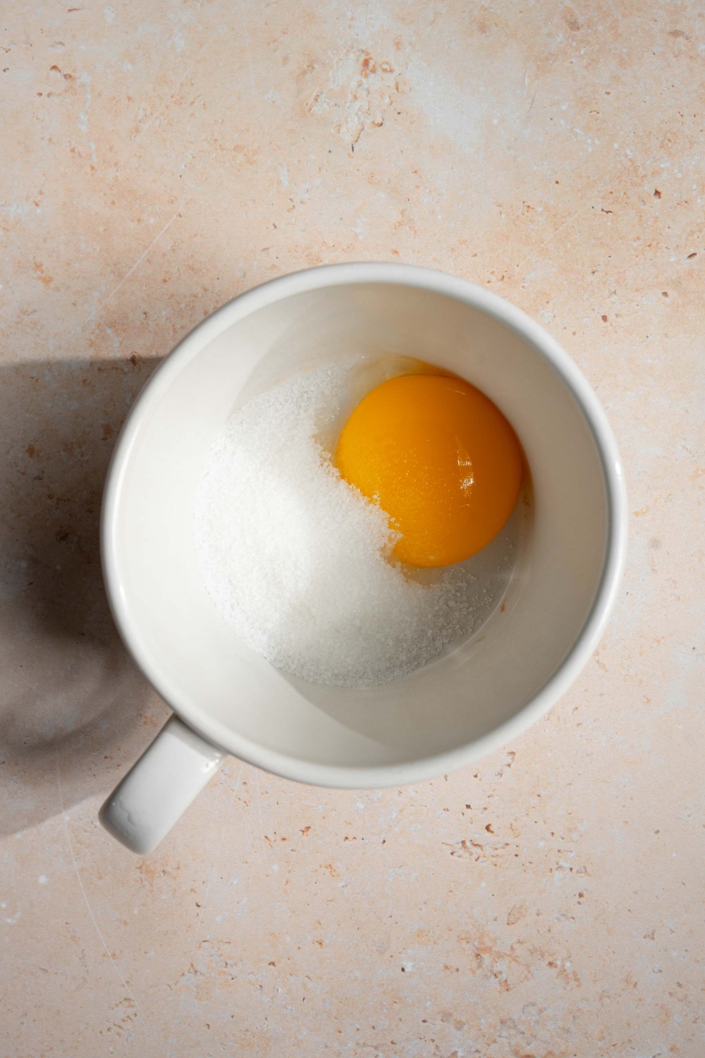 A white mug with sugar and an egg yolk. The mug is on a tan counter.