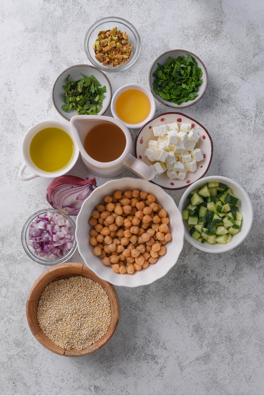 A marble counter with several bowls in various sizes containing ingredients to make Jennifer Aniston salad including quinoa, chick peas, cucumber, red onion, feta cheese, parsley, mint, roasted pistachios, lemon, honey, chicken stock, and more.