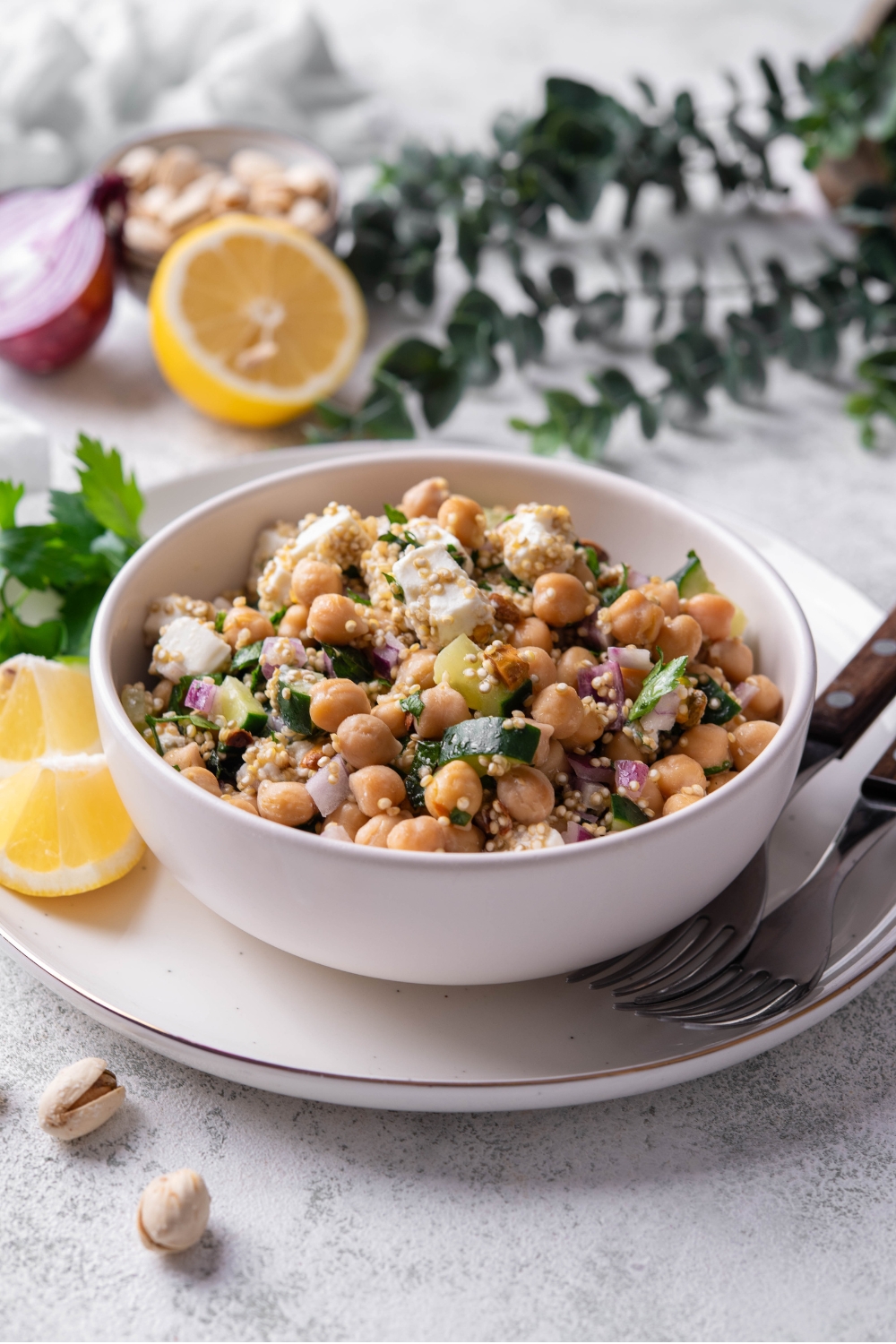A white bowl of Jennifer Aniston salad including chick peas, quinoa, cucumber, red onion, feta cheese, and pistachios tossed in a lemon dressing. The bowl is on a white plate with a fork and lemon. The plate is on a marble counter with lemon, onion, a bowl of chick peas, and a white cloth napkin.
