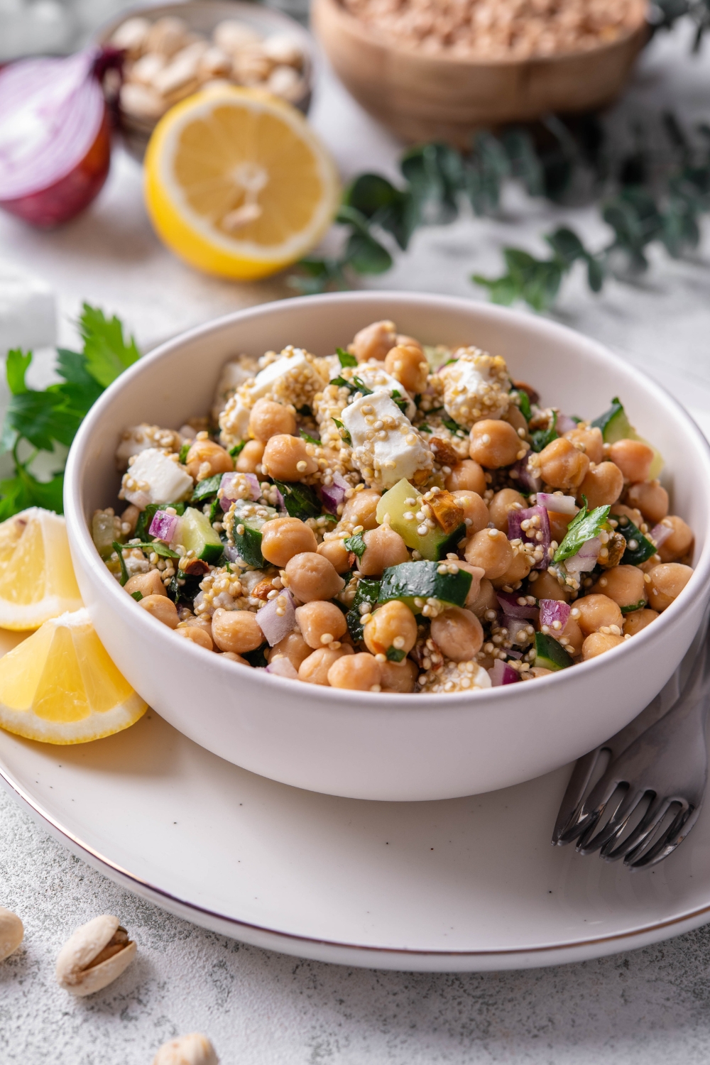 A white bowl of Jennifer Aniston salad including chick peas, quinoa, cucumber, red onion, feta cheese, and pistachios tossed in a lemon dressing. The bowl is on a white plate with a fork and lemon. The plate is on a marble counter with lemon, onion, a bowl of chick peas, and a white cloth napkin.