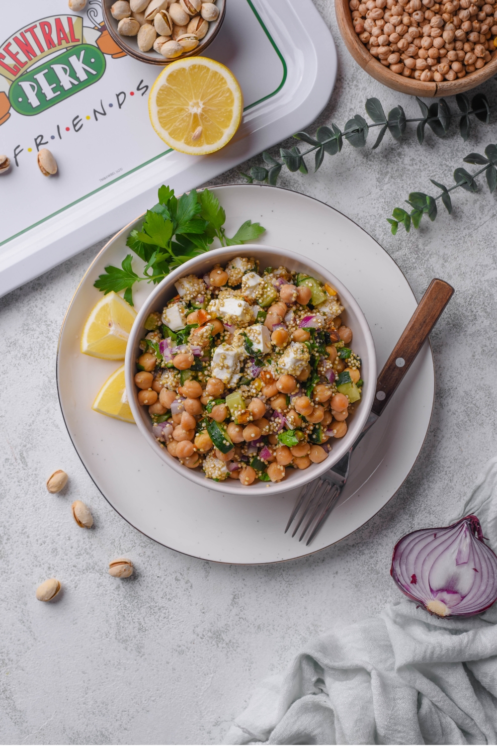 A white bowl of Jennifer Aniston salad including chick peas, quinoa, cucumber, red onion, feta cheese, and pistachios tossed in a lemon dressing. The bowl is on a white plate with a fork and lemon. The plate is on a marble counter with lemon, onion, a bowl of chick peas, and a white cloth napkin.