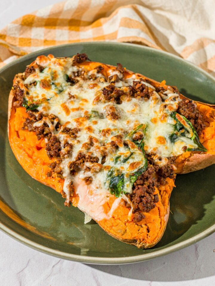 A green plate with a loaded sweet potato sliced open and stuffed with a beef mixture and topped with melted cheese. The plate is on a white counter with a yellow checkered napkin.