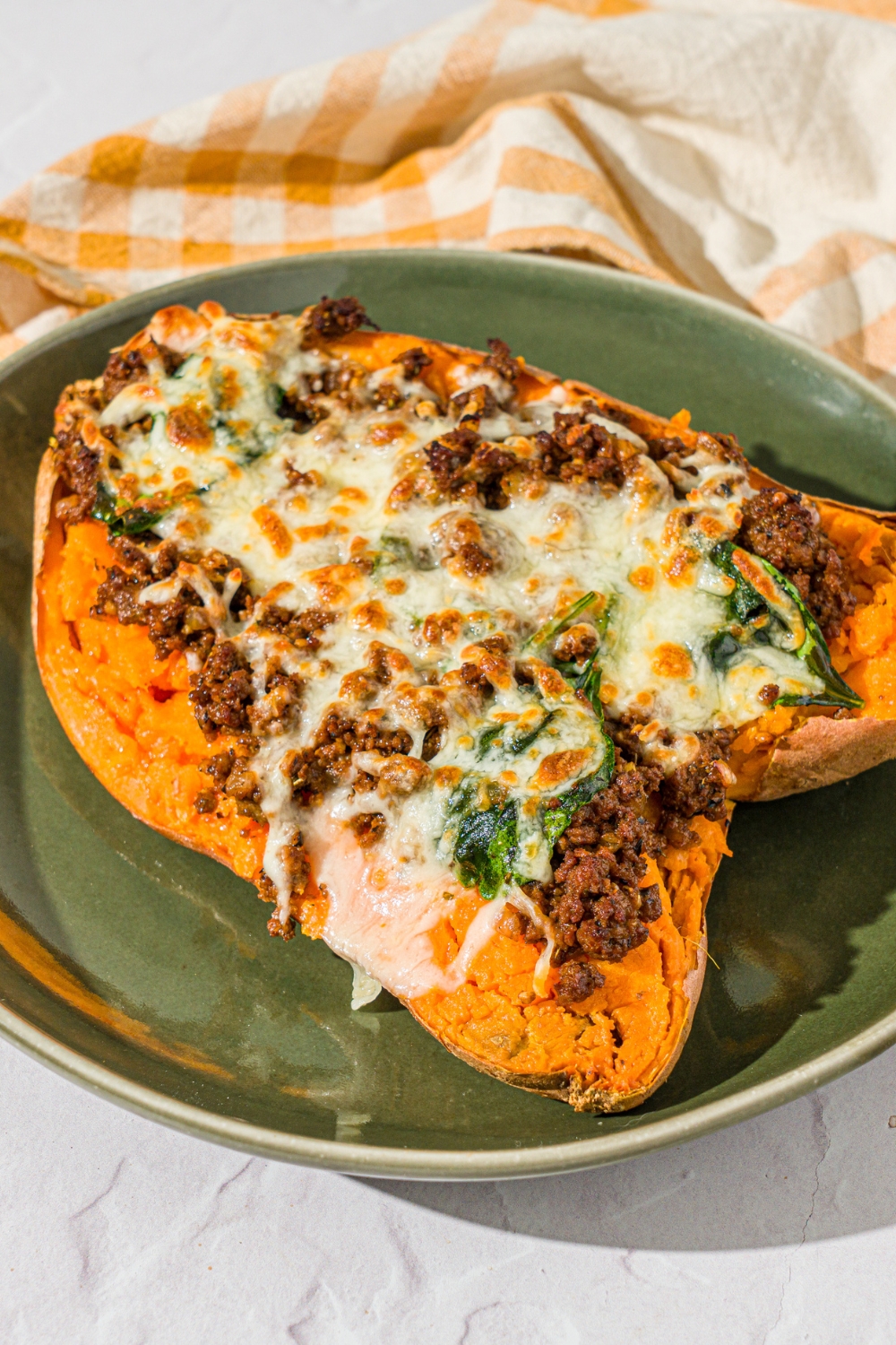 A green plate with a loaded sweet potato sliced open and stuffed with a beef mixture and topped with melted cheese. The plate is on a white counter with a yellow checkered napkin.