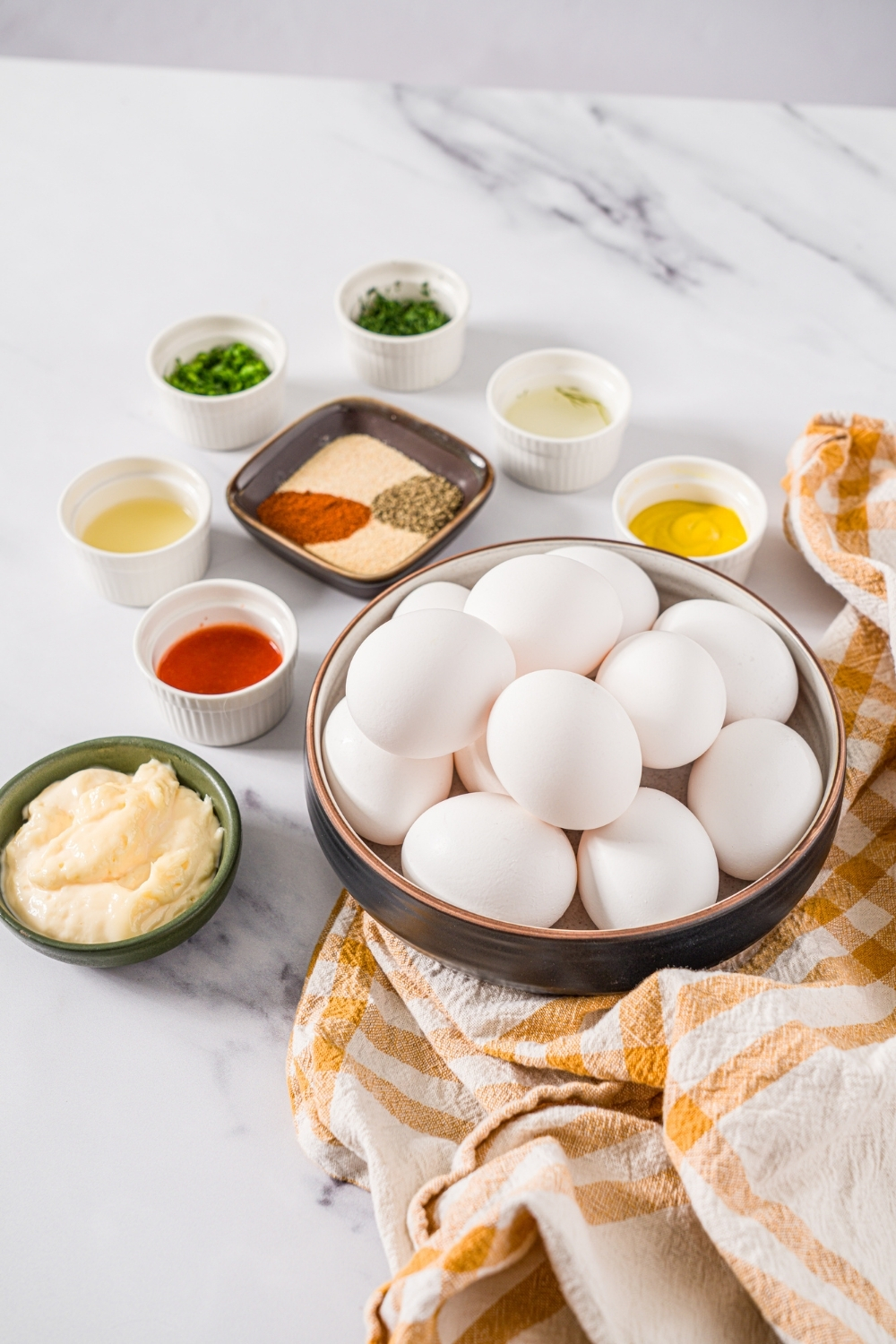 A marble counter with several bowls in various sizes containing ingredients to make Master's egg salad including eggs, mayo, mustard, dill, hot sauce, chives, and seasonings.