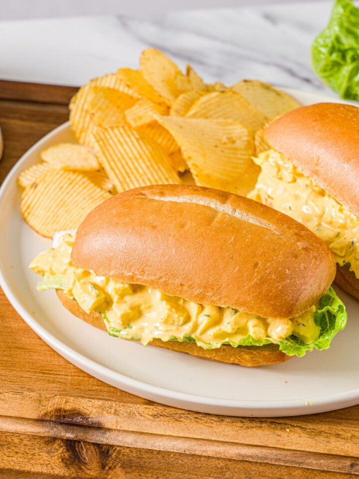 A white plate with two egg salad sandwiches on sandwich roll with lettuce. There plate is served with chips. The plate is on a wooden board.