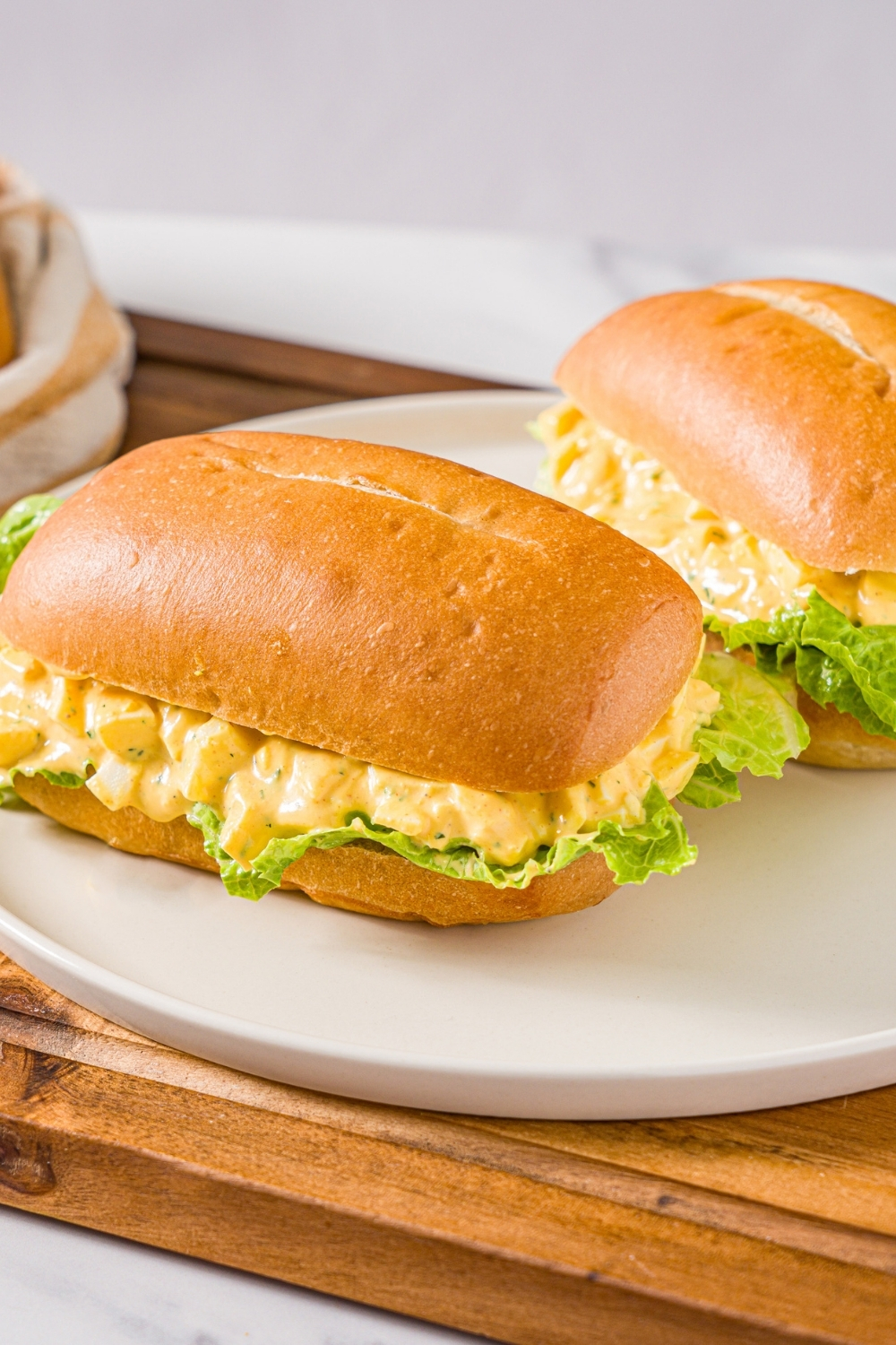 A white plate with two egg salad sandwiches on sandwich roll with lettuce. The plate is on a wooden board.