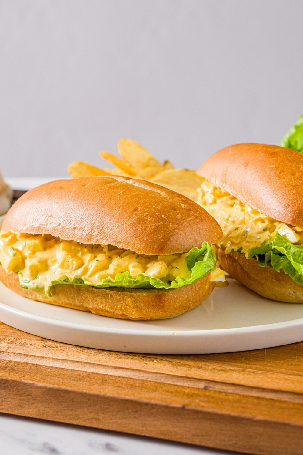 A white plate with two egg salad sandwiches on sandwich roll with lettuce. There plate is served with chips. The plate is on a wooden board.