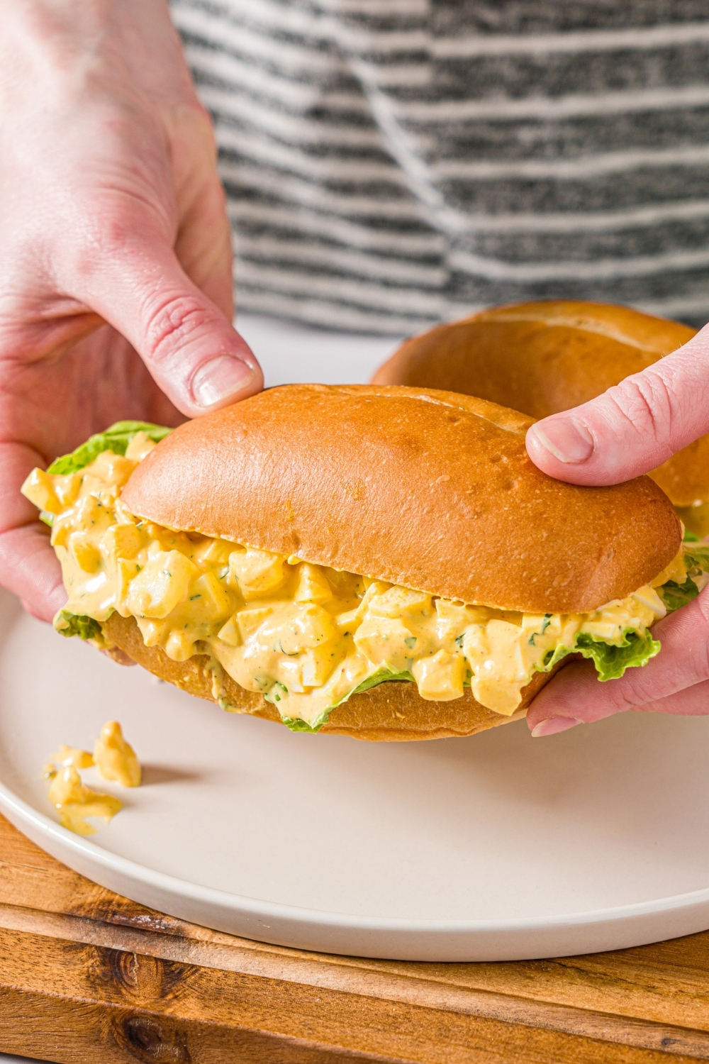 A hand holding an egg salad sandwich on a sandwich roll with lettuce over a white plate. There is an additional sandwich on the plate. The plate is on a wooden board.
