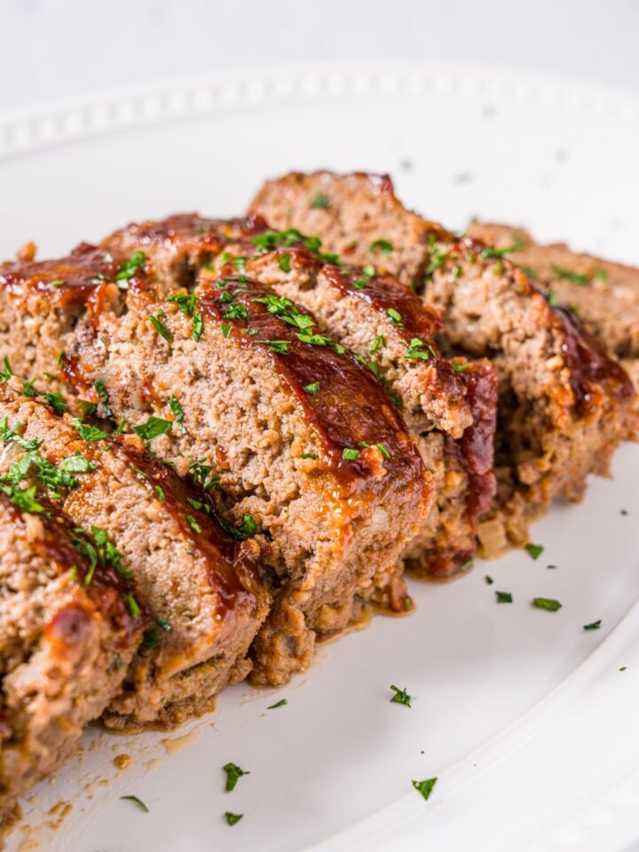 A white platter with sliced meatloaf topped with glaze and garnished with fresh parsley. The platter is on a marble counter.