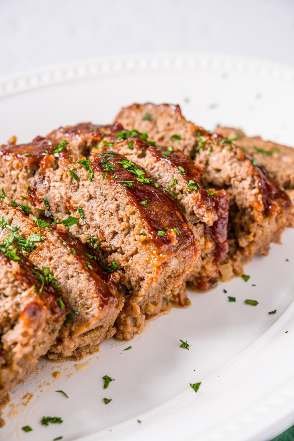 A white platter with sliced meatloaf topped with glaze and garnished with fresh parsley. The platter is on a marble counter.