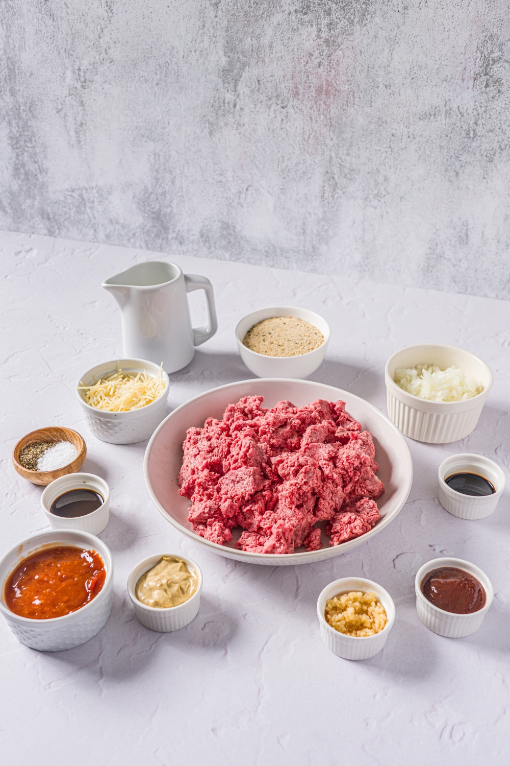 A marble counter with multiple bowls in various sizes containing ingredients to make meatloaf without eggs including ground beef, breadcrumbs, parmesan cheese, milk, diced onion, Worcestershire sauce, ketchup, mustard, and seasonings.